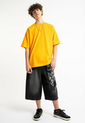 Nastolatek o kręconych włosach, ubrany w jaskrawożółtą, za dużą koszulkę, czarne długie spodenki z czarną bandaną i czarne trampki.