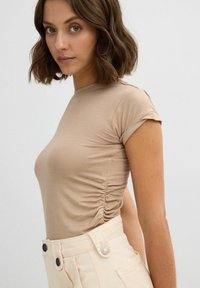 Camiseta ajustada de color beige con mangas cortas y fruncido lateral, combinada con pantalones de talle alto en un beige claro que presentan un detalle de botón.