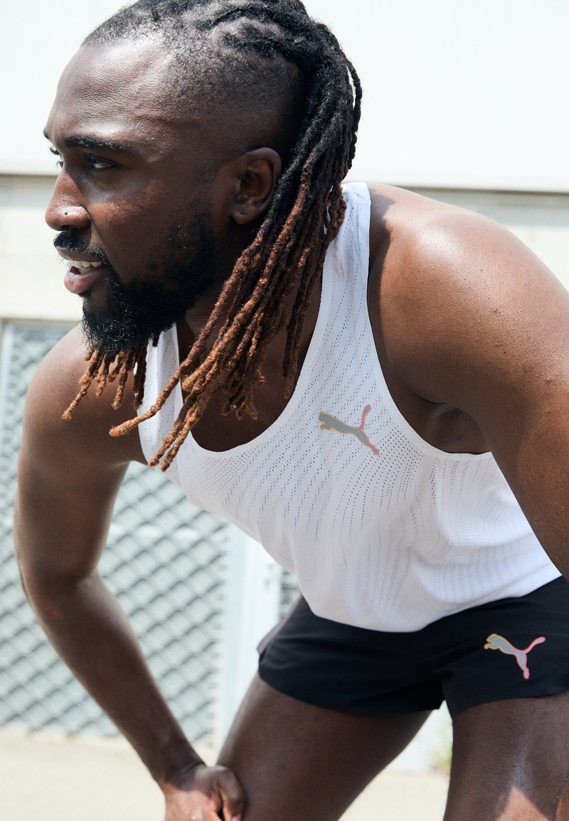 Canotta bianca in rete con loghi Puma dorati e rosa, abbinata a pantaloni corti neri con un piccolo logo Puma. Il soggetto si inclina in avanti, concentrato.