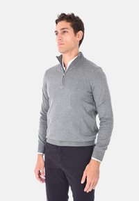 Maglione grigio a maglia con colletto con zip, orlo e polsini a coste, e una texture liscia, indossato sopra una camicia bianca. Pantaloni scuri completano l'outfit.