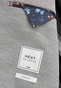 Szürke textúrájú szövet finom mintázattal. Virágmintás zsebbel és "NEXT TAILORING" és "SLIM FIT" feliratú címkékkel rendelkezik.