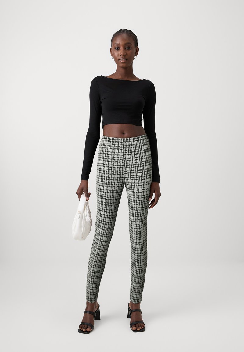 Svart långärmad cropped topp och gröna rutiga högmidjade leggings. Modellen håller en vit väska och bär svarta sandaler med remmar.