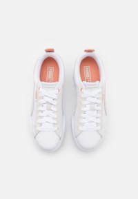 Hvite sneakers med beige aksenter, glatt tekstur, rund tå og snøring. Har en logo på tungen og myk innvendig polstring.