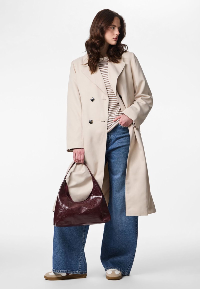 Frau mit beigem Trenchcoat, gestreiftem Hemd, Jeans mit weitem Bein, weißen Turnschuhen, hält eine dunkelrote Ledertasche, steht vor weißem Hintergrund.