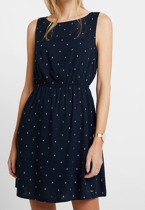 Ärmelloses marineblaues Kleid mit kleinen weißen Ankern, elastischem Taillenbund, getragen von einer Frau mit blondem Haar und Armbanduhr.