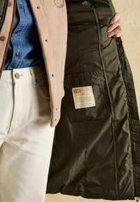 Szary polarowy kamizelka nałożona na niebieską dżinsową koszulę, odsłaniając zieloną pikowaną podszewkę z metką, oraz białe dopasowane spodnie z złotym guzikiem.