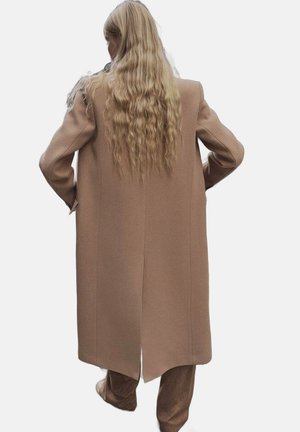Personne aux longs cheveux blonds ondulés portant un long manteau camel et un pantalon assorti, debout les mains sur les hanches, de dos.