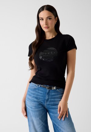 Kobieta z długimi ciemnymi włosami, ubrana w czarną koszulkę z logo Guess, niebieskie dżinsy i czarny pasek, stojąca na jasnym, jednolitym tle.