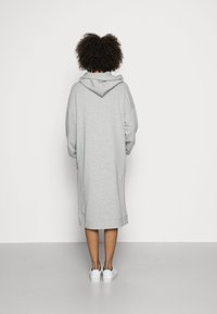 Persona con cabello rizado que lleva un vestido largo gris con capucha y zapatillas blancas, de pie frente a una pared blanca lisa.