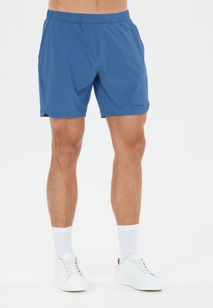 Man draagt blauwe sportshorts, witte enkelsokken en witte sneakers, staand tegen een effen achtergrond.