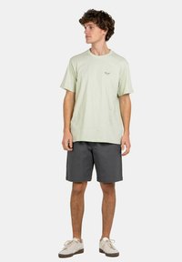 Hellgrünes Baumwoll-T-Shirt mit kurzen Ärmeln, natürliche Textur, ausgestattet mit einem kleinen Logo. Kombiniert mit dunkelgrauen Shorts und weißen Sneakers.