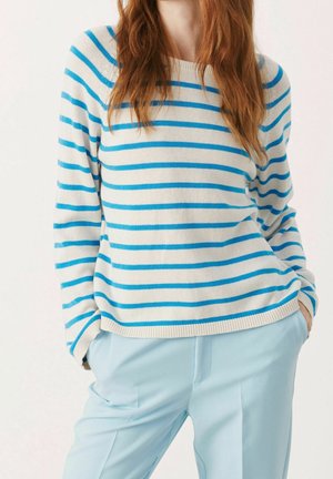 Kvinde iført en hvid sweater med blå vandrette striber og lyseblå bukser, hænderne i lommerne, står mod en ensfarvet baggrund.