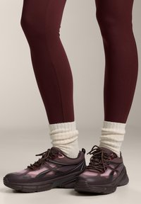 Bordó leggingset viselő lábak, krémszínű bordázott zoknik összegyűrve egy vastag sötétbarna és lila sportcipő felett, semleges háttéren.