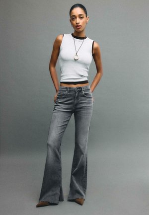 Donna in piedi con le mani nelle tasche posteriori, indossa una canotta bianca, jeans grigi svasati, scarpe marroni con punta a punta e una collana con pendente circolare.