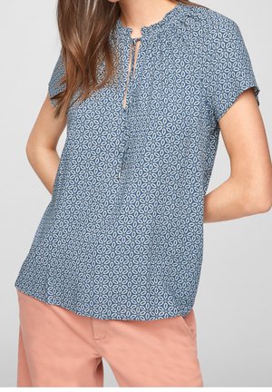 Woman wearing a blue patterned short-sleeve blouse with tie neckline and salmon-colored pants against a plain background.
