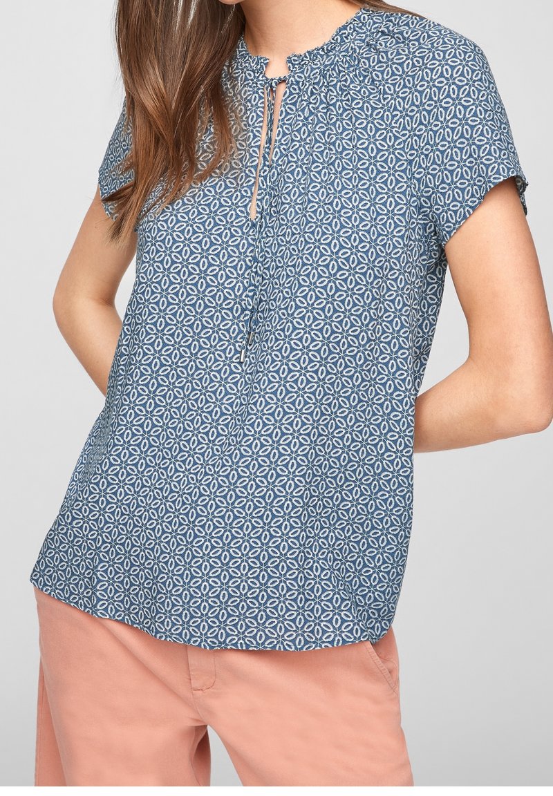 Femme vêtue d’un chemisier bleu à manches courtes à motifs avec un col à nouer et d’un pantalon couleur saumon sur un fond uni.