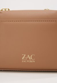 Borsa a tracolla in pelle color cuoio con dettagli in oro. Presenta una chiusura con zip e il logo impresso "ZAC ZAC POSEN" sulla parte frontale. Texture liscia.