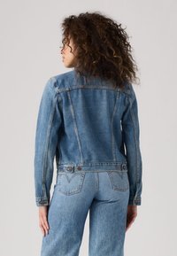 Giacca di jeans azzurro chiaro con taglio aderente, cuciture a contrasto e due bottoni sul retro. Presenta un colletto classico e tasche laterali.