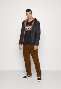Padded black jacket with an orange lining, worn over a black T-shirt featuring white text; paired with brown corduroy pants and black sneakers.