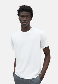 Camiseta blanca de algodón con cuello redondo, mangas cortas y un corte holgado. El modelo lleva pantalones cortos grises, mostrando un diseño sencillo.