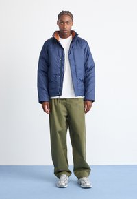 Navy puffer jacket with orange collar lining, zip closure, and elastic cuffs, paired with olive green pants and light gray sneakers.