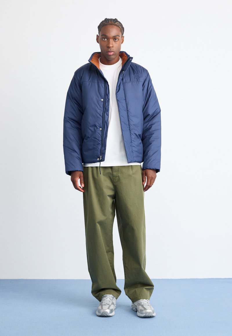 Giacca a vento navy con fodera arancione al colletto, chiusura con zip e polsini elasticizzati, abbinata a pantaloni verde oliva e sneaker grigio chiaro.