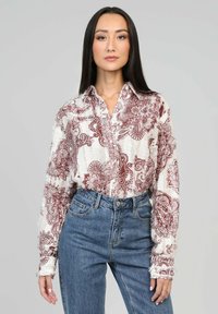Femme aux longs cheveux bruns portant une chemise blanche avec des motifs paisley rouges, rentrée dans un jean bleu taille haute, se tenant devant un fond uni.