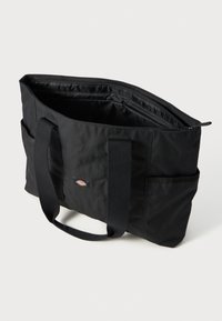 Svart stoff tote-veske med doble håndtak, glidelåslukking, utvendige lommer og en brodert logo. Innvendig er den romslig og uten fôr.