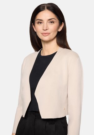 Frau mit glattem dunklem Haar trägt eine beige, kurz geschnittene Strickjacke mit offener Vorderseite über einem schwarzen Top und schwarzen Hosen vor einem schlichten hellen Hintergrund.