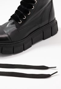 Botte en cuir noir avec semelle épaisse et lacets en velours, montrée avec deux lacets noirs lâches sur fond blanc.