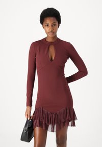 Burgundijas krāsu garām piedurknēm tērps ar atslēgvietas dekoltē un izplesti volāni apakšā no caurspīdīga, teksturēta auduma. Modelis rokās tur mazu melnu somiņu.