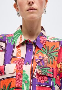 Γυναίκα με πολύχρωμο πουκάμισο με λουλουδάτα και πουλερικά μοτίβα, κουμπωμένο μπροστά, και μεγάλα ασημί σκουλαρίκια με αφηρημένο σχήμα.