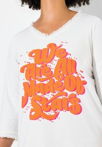 Witte shirt met korte mouwen en een versleten V-hals. Bevat feloranje en roze grafische tekst: "We Are All Made Of Stars," met accentpunten.