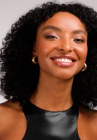 Sort ærmeløs top med en blank tekstur. Modellen har krøllet hår, bærer store gyldne øreringe i ringform og viser et lyst smil.