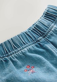 Modrá džínová látka s nařaseným elastickým pasem, zdobená vyšívaným květinovým motivem v červené a růžové barvě poblíž lemu.