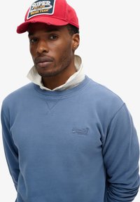 Lichtblauwe sweatshirt met een ronde hals, textuurstof en een klein logo. Gecombineerd met een rode pet met een patch.