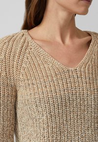 Vrouw in een beige en witte gebreide trui met een V-hals, die haar nek en een gedeeltelijk profiel toont.