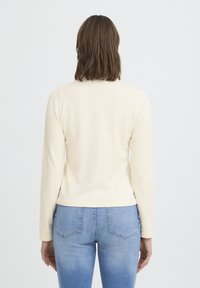 Haut à manches longues de couleur crème, fabriqué en tissu doux, avec un col rond et une silhouette ajustée, associé à un jean en denim bleu clair.