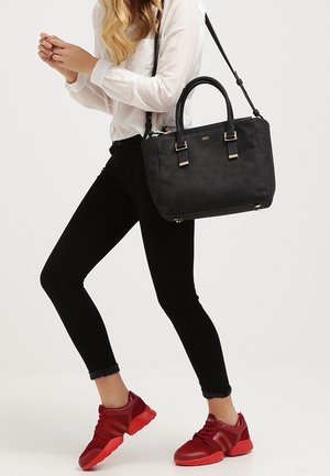 Mujer con pantalones negros y zapatillas rojas llevando un bolso negro con correa de hombro sobre fondo blanco.