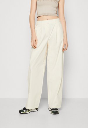 Pantalons larges créme avec des plis, en tissu lisse. Associés à des chaussures de sport noir et blanc.