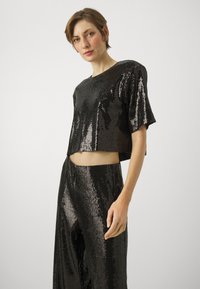 Svart paljett crop-top kombinert med matchende vide bukser. Jevn tekstur, glanset finish og korte ermer understreker moderne design.