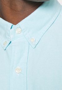 Světle modrá košilová košile s texturovanou tkaninou, s klasickým límečkem a perlovými knoflíky. Detailní záběr na límeček a horní část s knoflíky.