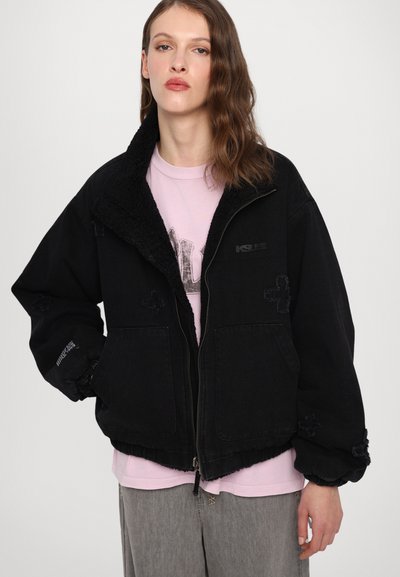 Black denim jacket with a soft, textured collar, zip front, elastic cuffs, and noticeable stitched accents. Worn over a light pink shirt.