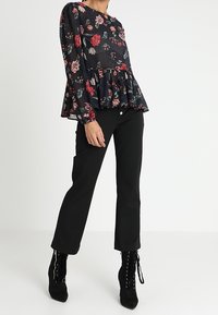 Blouse à peplum floral avec des manches longues sur fond sombre, ornée de fleurs rouges et blanches. Associée à un pantalon noir à jambes droites et à des bottines.