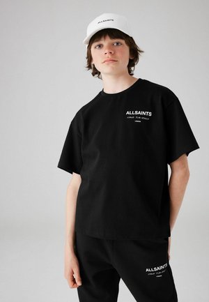 Camiseta de algodón negra de manga corta y escote redondo, con un logotipo blanco de "ALLSAINTS". Combinada con pantalones negros y una gorra blanca.