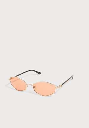 Gafas de sol con montura dorada, lentes geométricos tintados en rosa y puntas de patilla negras sobre fondo blanco.