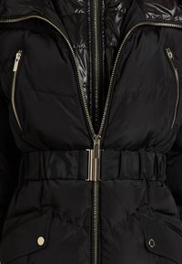 Czarna pikowana kurtka z błyszczącego materiału, podwójnymi zamkami, bocznymi kieszeniami zapinanymi na napy, paskiem w talii i złotymi akcentami metalowymi.