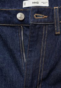 Jean en denim bleu foncé avec fermeture éclair avant partiellement ouverte, bouton en métal gravé "MNG JEANS ESTABLISHED 1994" et étiquette de marque visible.
