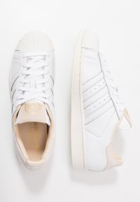 Zapatillas de cuero blancas con una puntera de goma texturizada, tres franjas plateadas y un logo dorado en la lengüeta. Diseño minimalista.