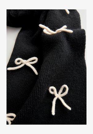 Mustan neulehuivin valkoisista langoista tehdyt solmukoristeet. Kankaan tekstuuri esittää pehmeää, kudottua kuviota, joka korostaa kontrastisia yksityiskohtia.